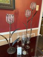Photo showing two metal candle holders with tulip-shaped glass shades, four assorted clear stemware glasses, and a David's Tea metal tea ball infuser on table.