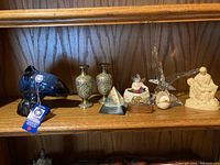 Wide view of shelf displaying assorted decorative items including a Blue Mountain dolphin, brass mini vases, glass bird, seated couple figurine, and trinkets.