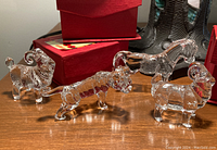 Four clear glass Chinese Zodiac animal figurines with intricate detail, including tiger, ram, horse, and goat/sheep, next to red gift boxes.