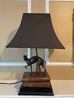 Full view of the greyhound desk lamp on a white surface showing the trapezoidal black fabric shade, bronze-tone greyhound figurine, and stacked vintage-style book base on black platform.