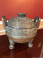 Front view image of the heavy, Asian inspired decorative ice bucket showing its ornate patina covered exterior with handles and tripod feet.
