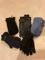 Five pairs of new mittens and gloves arranged on floor, showing variety of styles and colors including black leather gloves, blue fleece mittens, and insulated gloves.