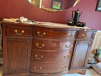 Wooden server with 6 drawers and 2 cabinets, brass handles, curved front, brown finish, polished surface, placed in a red wall room with mirror above.