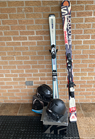 Full lot showing two pairs of skis and three helmets on a floor mat with brick wall background.
