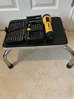 Black step stool with metal legs holding screwdriver set and flashlight