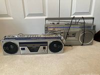 Photo shows two vintage portable radio cassette stereo units placed on carpet: one Toshiba with dual prominent circular speakers and cassette deck, and one JVC with large single speaker, cassette deck, and radio tuner across top.