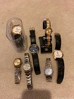 Photo showing seven wristwatches of different brands including Seiko, Citizen, Timex arranged on carpet with one watch on black display stand and another inside plastic tube container.