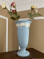 Light blue Wedgwood vase with six ceramic floral stems arranged inside, placed on a wooden surface in a neutral room with crown molding visible.