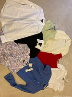 Photo showing eight women's tops on hangers spread out on a surface, including Ralph Lauren blue button-down shirt with tag, a black top with white collar, cream zip-up sweater, light green top, red sleeveless top, floral blouse, blue button-down shirt, and a white shirt.