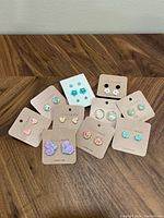 Top down view of earrings arranged on multiple studioxviii cards on a wooden surface. Various designs including flowers, hearts, hexagons, and circular studs are visible.