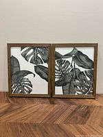 Two framed pictures with black and white tropical leaf patterns standing side by side on wooden surface against neutral wall.