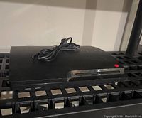 Sony PS3 Slim console with power cable placed on top, showing front disc slot and embossed PS3 logo on console top surface.