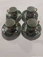 Set of four china cups and saucers arranged on white surface, decorated with hummingbird and floral pattern with green and gold accents.