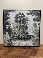 Front view of square framed canvas wall art depicting black and white tropical foliage and trees.