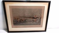 Framed equestrian print showing horses and riders in red hunting jackets jumping over water and fences in a naturalistic scene.