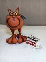 Front view of orange Creatures of Delight bat sculpture with large feet and expressive eyes on white surface with attached tags.