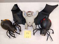 Front view of the gray bat in center with two black spiders on either side on white background with yellow note showing size