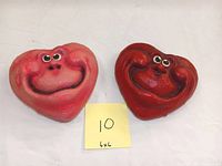 Front view of two anthropomorphic heart sculptures, one pink and one red, both have smiling faces and bulging eyes.