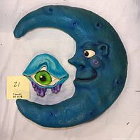 Photo showing the blue crescent moon sculpture with human face and the separate blue eye sculpture with green iris and purple drips from a top angle on a white background with a note indicating dimensions 15x16.