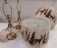 Pair of Golden Lipper metal lamp bases with harp fittings alongside two decorative drum lampshades featuring deer and forestry silhouettes. Lamps and shades are displayed separately on a white surface.