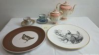 Two decorative porcelain plates with bird illustrations beside a Coalport teapot, creamer, and two demitasse cups with saucers on white cloth.