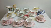 Top view of ten assorted cups and saucers with various styles including floral, gold trim, and pink geometric patterns.