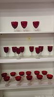 A shelf holding three medium-sized ruby red stem glasses on the top row, nine smaller ruby red stem glasses in the middle, and five wider bowl-shaped ruby red stem glasses on the bottom.