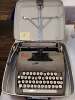 Vintage Smith-Corona manual typewriter inside open metal carrying case, with visible handwriting on typewriter body.