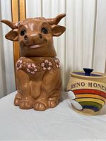Photo showing the ceramic cow cookie jar with pink flower decorations and the Reno Money ceramic jar with rainbow design side by side on a table.