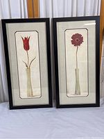Two vertical framed floral prints, each featuring a single flower in a narrow vase, with black frames and glass, placed side by side.