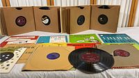 Vintage 9 1/2 inch vinyl records displayed with paper sleeves, brown cardboard storage boxes, and colorful paper inserts showing musical notes and advertisements.