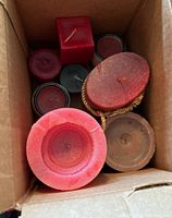 Box containing assorted candles of various sizes, shapes (round, oval, square), and colors including red, brown, and black, some in metal tins.