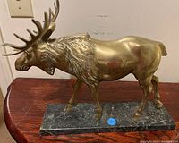 Side view of brass moose ornament showing full body and green marble base on wooden table against white wall.