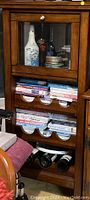 Front view of the wooden cabinet showing the glass door with a round silver handle, and three curved shelves holding wine bottles and books.