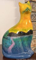 Photo of ceramic cat ornament showing the colorful mountain scene painted on its body, with yellow, green, blue, black, and pink colors visible.