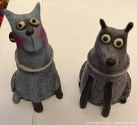 Photo showing two small ceramic dog statues with cartoon-like features, one bluish and one purplish, sitting side by side on a white surface.