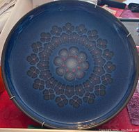 Top down view of a blue circular decorative plate with a detailed and intricate geometric floral pattern with red dot accents. Plate is glossy and mounted on a silver metal hanger.