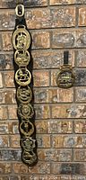 Photo of large and small decorative horse brass mounted on black leather straps hanging against a brick wall.