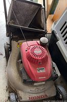 Angled view of mower showing red engine cover, aluminum deck and attached mesh grass bag