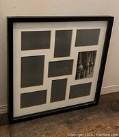 Photo of black wooden multi-opening photo frame with white mat and eight photo slots, leaning against a wall.