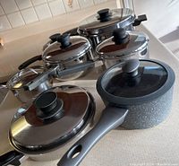 Shows the full set of 8 saucepans with lids, including a speckled non-stick pan with glass lid and mostly stainless steel saucepans with metal lids and black handles.