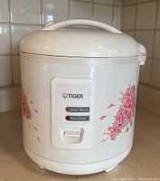 Closed rice cooker showing white body with pink floral rose decorations and front control panel with Keep Warm and Rice Cook buttons.
