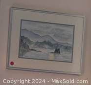 Framed watercolour painting depicting a coastal landscape with a boat, rocky shore, and mountains in the background. The frame is light-colored with a white matting.