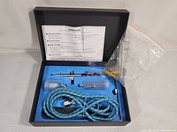 Photo showing contents of the open airbrush kit box with airbrush gun, hoses, fluid cups, and instruction sheet visible.