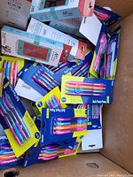 Multiple packs of colorful retractable ballpoint pens and boxed kids blue light glasses in cardboard box.