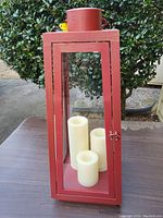 Antique red tapered lantern with three cream-colored flameless candles inside, sitting outdoors on a table.