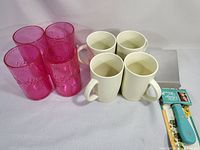 Four translucent pink drinking cups, four white Starbucks coffee mugs, and a Pioneer Woman spatula in original packaging on a white cloth background.