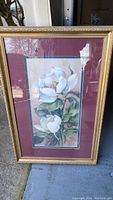 Full view of signed framed and matted magnolia floral artwork in gold frame with glass on concrete floor.