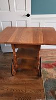 Side view of the wooden tea cart showing drop leaf extended, large wooden decorative wheel and caster wheel on turned legs.