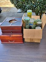 Photo showing two wood jewelry boxes stacked beside a cardboard box containing multiple glass bottles outdoors.